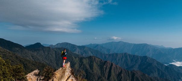 Hiker stands atop a rocky cliff overlooking expansive mountain range in Vietnam.